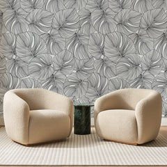 Papier peint panoramique Monstera au Trait Noir, posé sur toute la paroi d'un salon, derrière deux fauteuils en bouclette crème et un guéridon en marbre vert