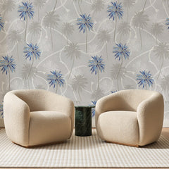 Papier peint panoramique Palmeraie Bleue sur Gris, posé sur toute la paroi d'un salon, derrière deux fauteuils en bouclette crème et un guéridon en marbre vert