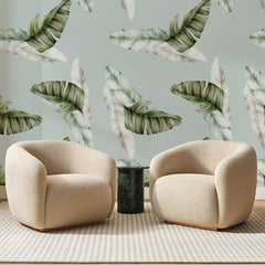 Papier peint panoramique Feuilles de Bananier Sauge, posé sur toute la paroi d'un salon, derrière deux fauteuils en bouclette crème et un guéridon en marbre vert