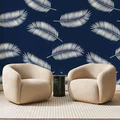 Papier peint panoramique Palmes Blanches sur Bleu Marine, posé sur toute la paroi d'un salon, derrière deux fauteuils en bouclette crème et un guéridon en marbre vert