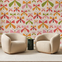 Papier peint panoramique Papillons Graphiques Corail, posé sur toute la paroi d'un salon, derrière deux fauteuils en bouclette crème et un guéridon en marbre vert