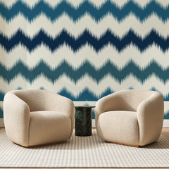 Papier peint panoramique Chevrons Ikat Bleu Nuit, posé sur toute la paroi d'un salon, derrière deux fauteuils en bouclette crème et un guéridon en marbre vert
