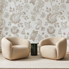 Papier peint panoramique Cachemires et Colibris Taupe, posé sur toute la paroi d'un salon, derrière deux fauteuils en bouclette crème et un guéridon en marbre vert.