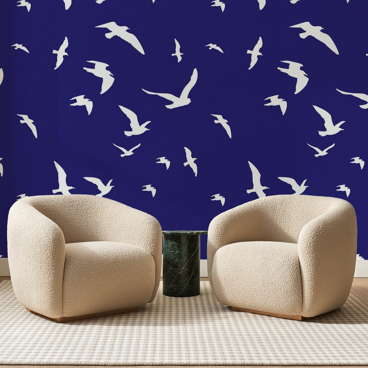 Papier peint panoramique Mouettes Blanches sur Outremer, posé sur toute la paroi d'un salon, derrière deux fauteuils en bouclette crème et un guéridon en marbre vert