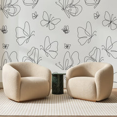 Papier peint panoramique Papillons au Trait Noir, posé sur toute la paroi d'un salon, derrière deux fauteuils en bouclette crème et un guéridon en marbre vert