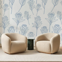Papier peint panoramique Protéas au Trait Bleu, posé sur toute la paroi d'un salon, derrière deux fauteuils en bouclette crème et un guéridon en marbre vert