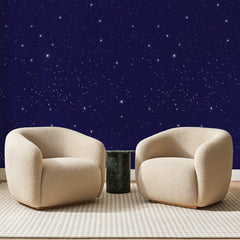 Papier peint panoramique Ciel Étoilé Bleu Nuit, posé sur toute la paroi d'un salon, derrière deux fauteuils en bouclette crème et un guéridon en marbre vert